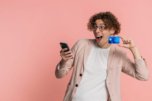 Happy Caucasian young man with curly hair using credit bank card and smartphone while transferring spending money, purchases online shopping, ordering food delivery. Guy isolated on pink background