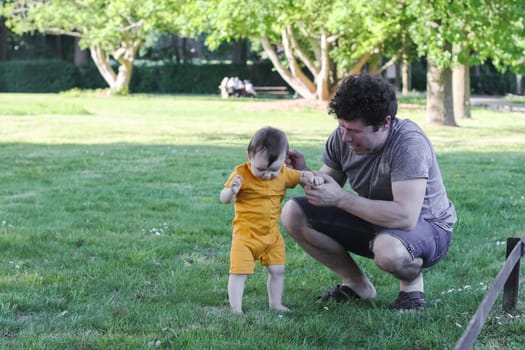 Caucasian young dad in a blue t-shirt with curly brown hair teaches how to walk and take the first steps to his little daughter in a yellow bodysuit, standing barefoot on the grass and looking at a daisy in a public park, close-up side view. The concept of fatherhood, dads, family vacation, spring walk, first steps, happy childhood, parks and rec.
