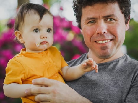 Caucasian young dad man in a gray t-shirt with a happy smile hugs his little daughter in a yellow bodysuit with a daisy in her mouth in a public park, close-up side view. Concept of fatherhood, dads, family vacation, lesson, spring walk, first steps, dad care .