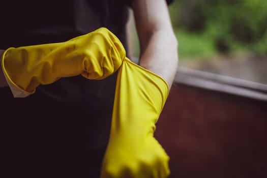 One young caucasian unrecognizable male builder in black uniform clothes pulling on yellow rubber gloves stretching his hand forward while standing near the window, close-up view from below. The concept of home renovation, construction work.