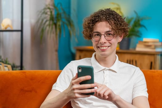 Caucasian young man sitting on couch using mobile phone smiles at home living room apartment. Happy guy with curly hair texting share messages content on smartphone social media applications online.