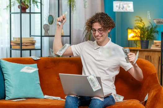 Money cash dollar rain falling on Caucasian man working on laptop computer celebrating success, winning, goal achievement, good news lottery jackpot luck. Career grow. Young guy at home room on sofa.