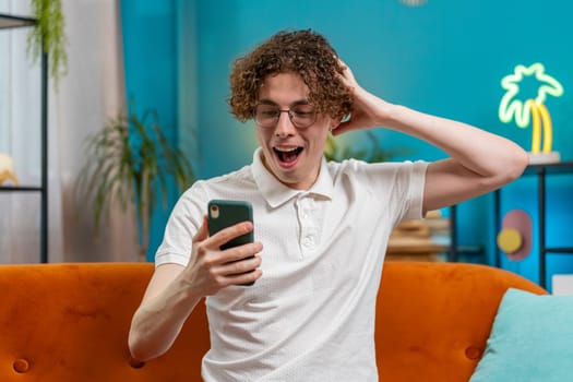 Surprised excited Caucasian young man winner holding smartphone reading good news amazed by online bet bid game win. Happy guy male on sofa looking at screen overjoyed by victory success in apartment.