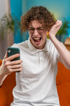 Sad displeased Caucasian man using smartphone typing browsing, loses becoming surprised sudden lottery game results, bad news, fortune loss, fail, deadline, virus. Young guy with curly hair at home.