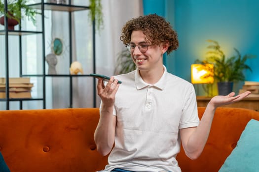 Happy Caucasian man making phone call conversation with friends at modern home room apartment. Young brunette guy enjoying mobile loudspeaker talking gossip, rumors, good news sitting on couch indoors