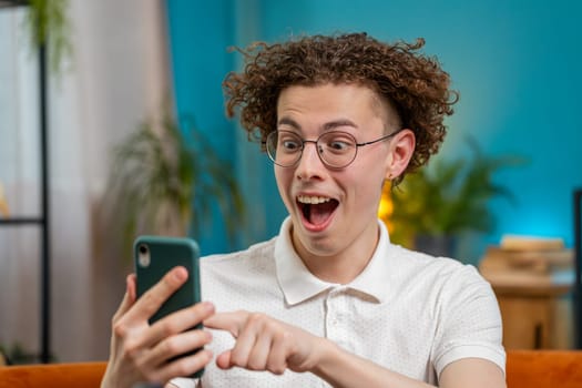 Surprised excited Caucasian young man winner holding smartphone reading good news amazed by online bet bid game win. Happy guy male on sofa looking at screen overjoyed by victory success in apartment.