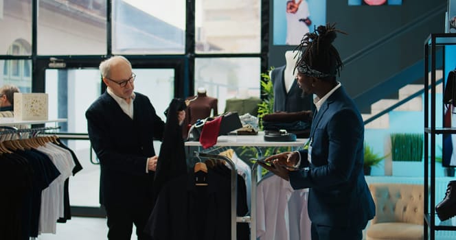 African american employee giving advice to senior customer in retail store, trying to find an elegant shirt on sale. Elderly client asking assistant for suggestions and recommendations. Camera B.