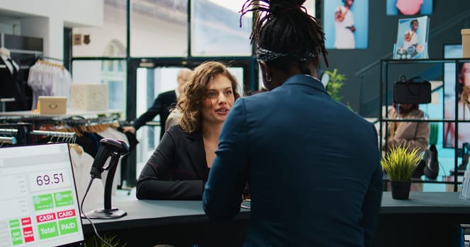 Trendy woman prepared to buy designer bag at store checkout, choosing modern accessories on sale. African american employee at cash register selling items to regular customer. Camera B.