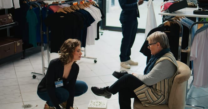 Elderly woman searching for a comfortable pair of shoes at mall, asking retail assistant about available sizes. Store employee recommending various footwear models for senior client. Camera B.