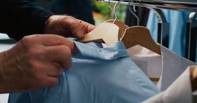 Elderly client checking all clothes placed on display at showroom, browsing through hangers to find items on discount. Senior man shopping for elegant shirts and suit jackets. Close up. Camera B.