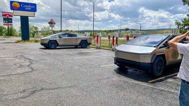 Las Vegas, New Mexico, USA-June 2, 2024-Two Tesla Cybertrucks parked and charging at an outdoor Supercharger station on a cloudy day. The station is surrounded by trees and open land, creating a scenic backdrop for the futuristic vehicles.