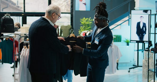 African american store assistant helping regular client with a suit in shopping center, senior customer looking to buy formal clothes. Elderly man shopping for new collection at the mall. Camera B.