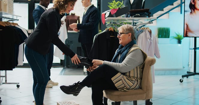 Retail worker presenting various pairs of shoes for senior client, assisting woman to find the perfect footwear in fashion showroom. Elderly customer trying on footgear before buying. Camera B.