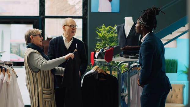 Store employee helping senior people with clothing suggestions, presenting new fashion collection items to couple in department store. African american employee makes recommendations. Camera A.