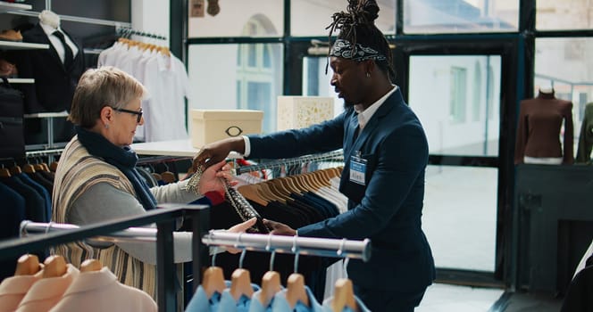 Elderly client asking to see multiple trendy bags in clothing store, talking to shop assistant about recommendations. African american worker shows different designer accessories. Camera B.