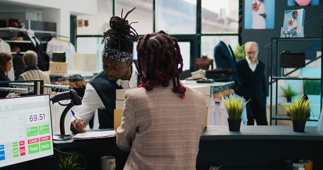 African american guy picking up order from clothing store, signing confirmation papers at cash register. Woman employee preparing boxes with requested fashion items and accessories. Camera B.