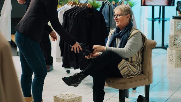 Retail worker presenting various pairs of shoes for senior client, assisting woman to find the perfect footwear in fashion showroom. Elderly customer trying on footgear before buying. Camera A.