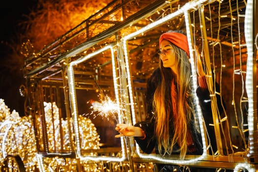 Woman holding sparkler night while celebrating Christmas outside. Dressed in a fur coat and a red headband. Blurred christmas decorations in the background. Selective focus.