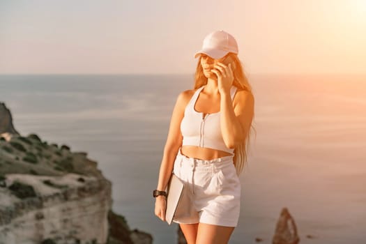 A happy woman in white shorts and T-shirt enjoys the picturesque sea view while holding a laptop and talking on the phone