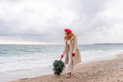 Redhead woman Christmas tree sea. Christmas portrait of a happy redhead woman walking along the beach and holding a Christmas tree in her hands. She is dressed in a light coat and a red beret