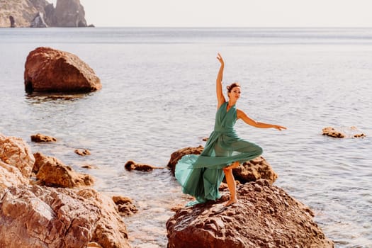 Woman green dress sea. Female dancer in a long mint dress posing on a beach with rocks on sunny day. Girl on the nature on blue sky background