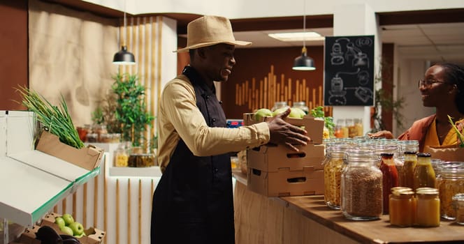 Local vendor placing crates with freshly harvested fruits on grocery store display, recommending organic produce to regular customer. African american farmer restocking all merchandise. Camera 2.