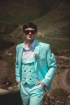 Front view of handsome groom in stylish wedding suit with white shirt and tie standing on hill and forested mountains looking at camera.