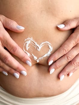 Close up of womans hands middle age with white nails applying cream in the shape of heart on her stomach. Skincare, self care, and personal hygiene with on love and body positivity. High quality photo