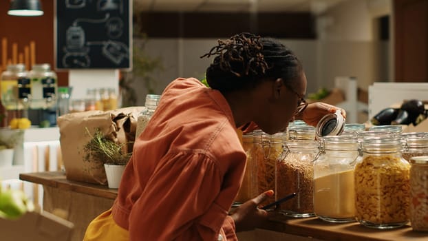 Customer checking shopping list on phone before choosing products, trying to buy organic locally grown food for low carbon footprint. African american woman looking at checklist notes. Camera 1.