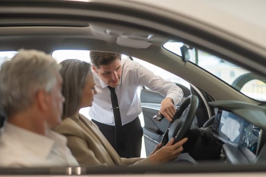 A gray-haired Caucasian woman is sitting behind the wheel of a car. Car dealership salesman consulting mature married couple