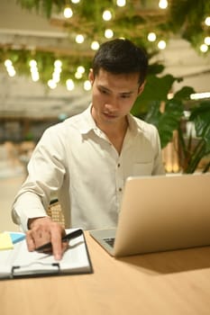 Millennial male using laptop and working with financial document in modern office lounge.