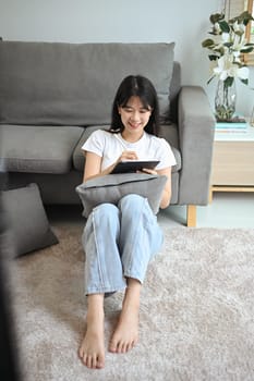 Charing young female freelancer using digital tablet sitting on floor in living room.