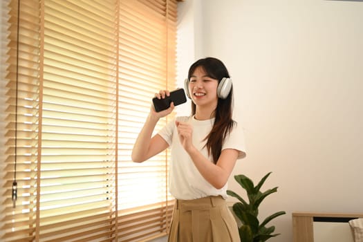 Happy young asian woman having fun listening to music and singing in living room.