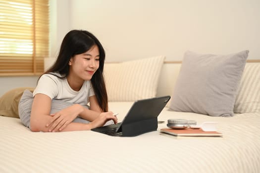 Smiling young woman in casual clothes using digital tablet while laying on bed.
