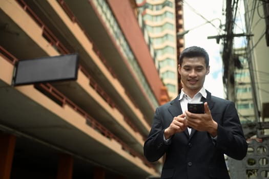 Handsome businessman in formal wear walking on city street and using smartphone.