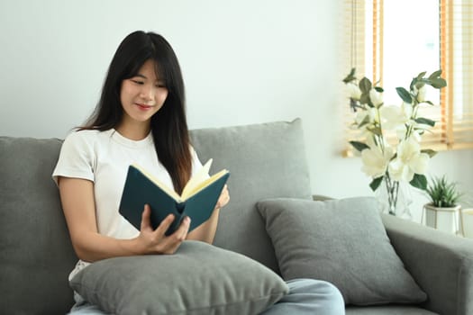 Young Asian woman spending leisure time at home reading a book sitting on couch.