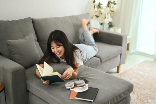 Happy young Asian woman reading book while lying on couch at home.