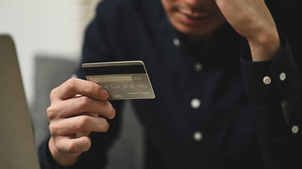 Upset young man holding credit card experiencing problems during payment online, overspending.