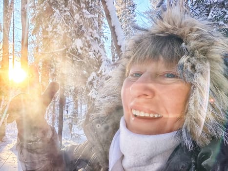 A cheerful middle aged woman in a winter coat with fur, scarf taking selfie outdoors on nature in a forest or park in beautiful sunny day