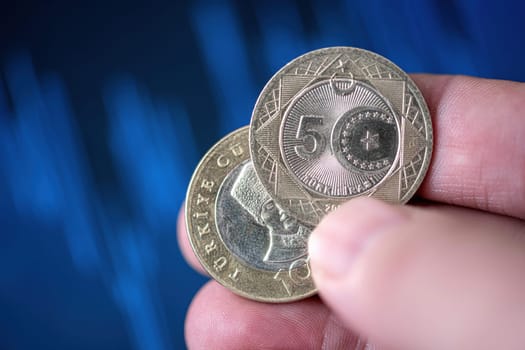 Hand holding a 5 Turkish Lira coin in front of a screen with financial chart curves