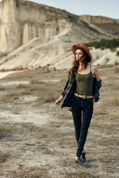 Serene woman in hat and green shirt standing with hands in pockets in front of majestic mountain