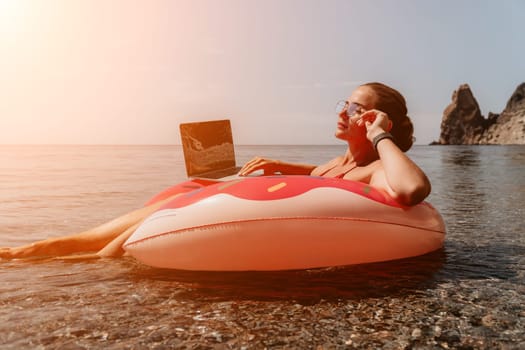 Woman freelancer works on laptop swimming in sea on pink inflatable ring. Pretty lady typing on computer while floating in the sea on inflatable donut at sunset. Freelance, remote work on vacation