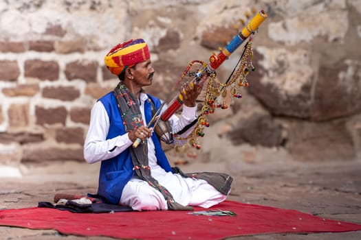 Jodhpur, Rajasthan, India - 26th Dec 2023: Rajasthani Indian musician sitting cross legged in colorful turban and white kurta pyjama local clothing in front of a brick wall at a mahal palace