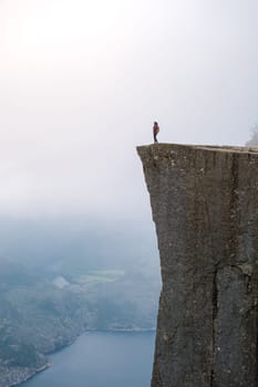 Preikestolen, Norway, A lone hiker stands on the edge of a dramatic cliff in Norway, overlooking a misty valley and a winding fjord.