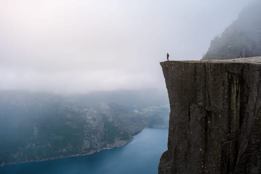 A lone hiker stands on the edge of Preikestolen, a dramatic cliff overlooking a misty fjord in Norway. Preikestolen Norway