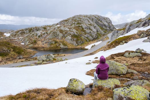 A lone hiker sits on a rock, gazing out at a snow-covered mountain pass in Norway, with a winding road and a small lake in the distance.