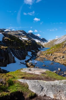 A winding river flows through a scenic valley in the Norwegian mountains, surrounded by snow-capped peaks and a clear blue sky. Kjeragbolten Norway