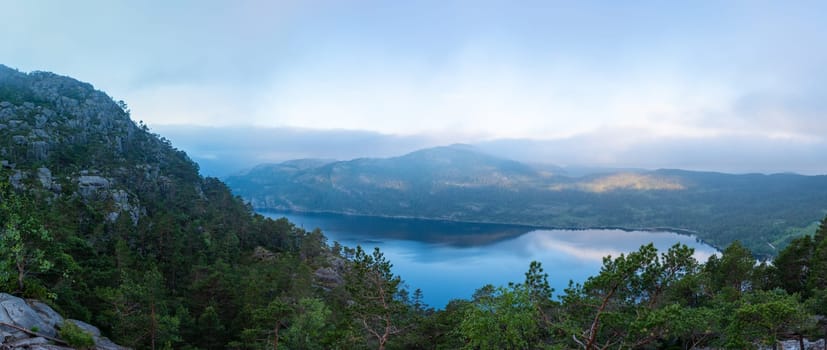 A panoramic view of a lake nestled amongst rolling hills in Norway, with a hazy mist settling over the landscape. Preikestolen, Norway
