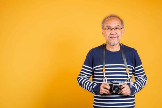 Portrait Asian old man wearing glasses holding vintage SLR camera studio shot isolated yellow background, smiling happy Photographer elderly man gray haired holds old fashioned camera