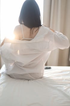 Young woman in lingerie and white shirt sit on bed indoors at home stretching in the morning after waking up, back view. Relaxing , Cozy, good morning, comfort, intimacy concept.